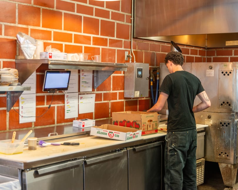 A chef in a black shirt and jeans unpacks tomatoes in a commercial kitchen with brick walls.