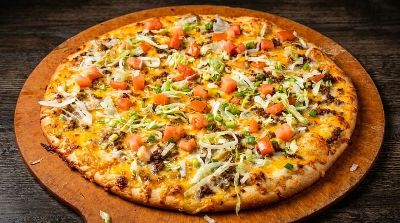 Taco pizza with ground beef, shredded lettuce, diced tomatoes, and cheese on a wooden board.