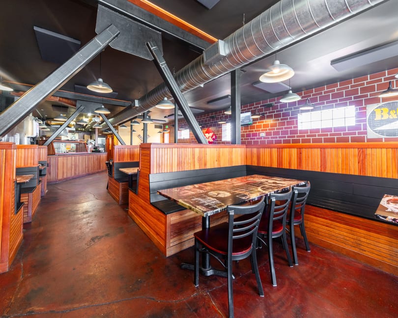 Restaurant interior with wooden booths, exposed brick, industrial ceiling, and a long counter in the background.