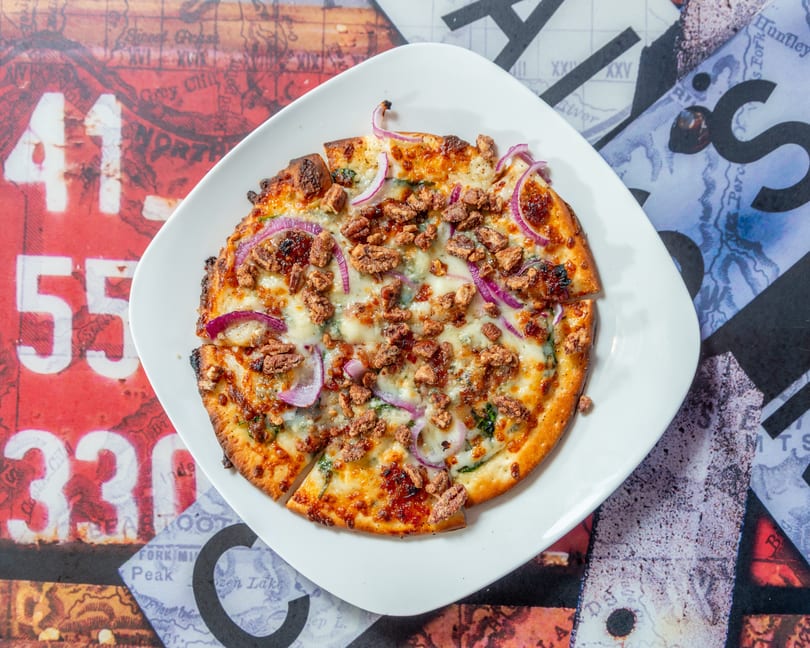 A round flatbread pizza with cheese, red onions, and pecans, on a white plate over a colorful map.