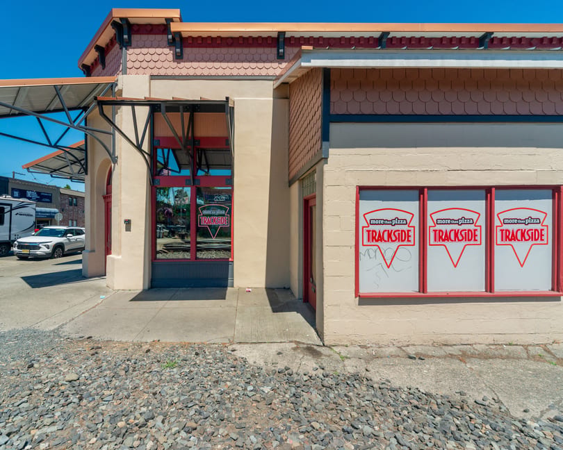 Exterior of Trackside Pizza restaurant with red-framed windows, a beige facade, and a gravel foreground.