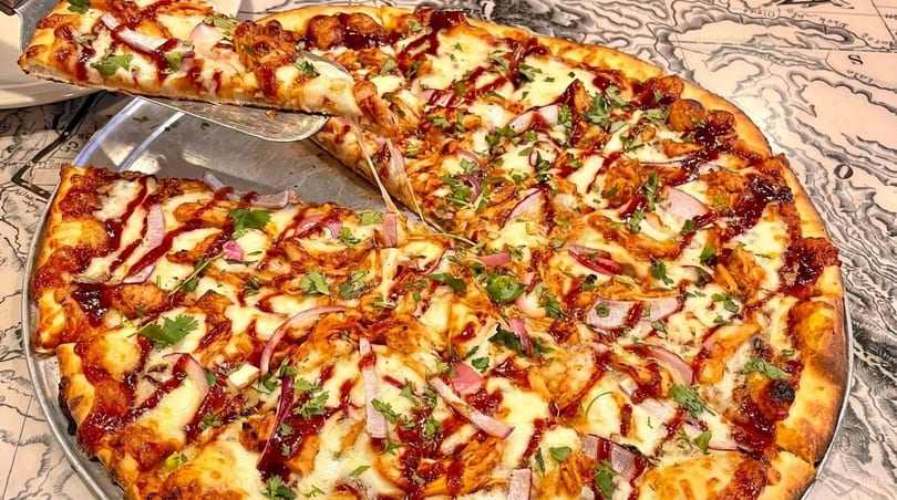 A slice of BBQ chicken pizza with red onions and cilantro is lifted from a whole pizza.